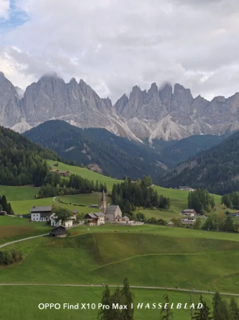 Petit village de montagne avec une église entouré de prairies verdoyantes sous une chaîne de montagnes aux sommets escarpés.