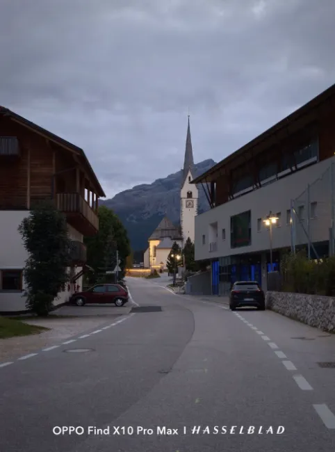 Rue d'un village alpin au crépuscule avec une église à tour pointue en arrière-plan sous un ciel nuageux.