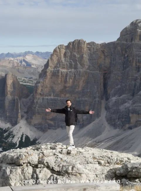 Homme posant à bras ouverts au sommet d'une montagne rocheuse devant de grands massifs de pierre.