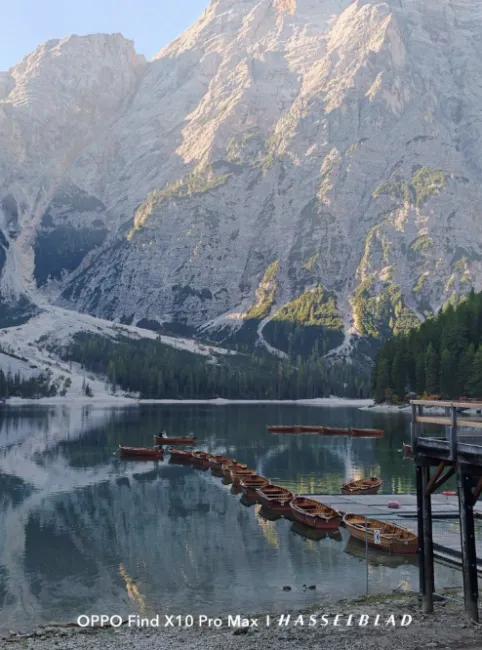 Des rangées de bateaux en bois sur un lac alpin reflétant la face illuminée d'une montagne rocheuse.