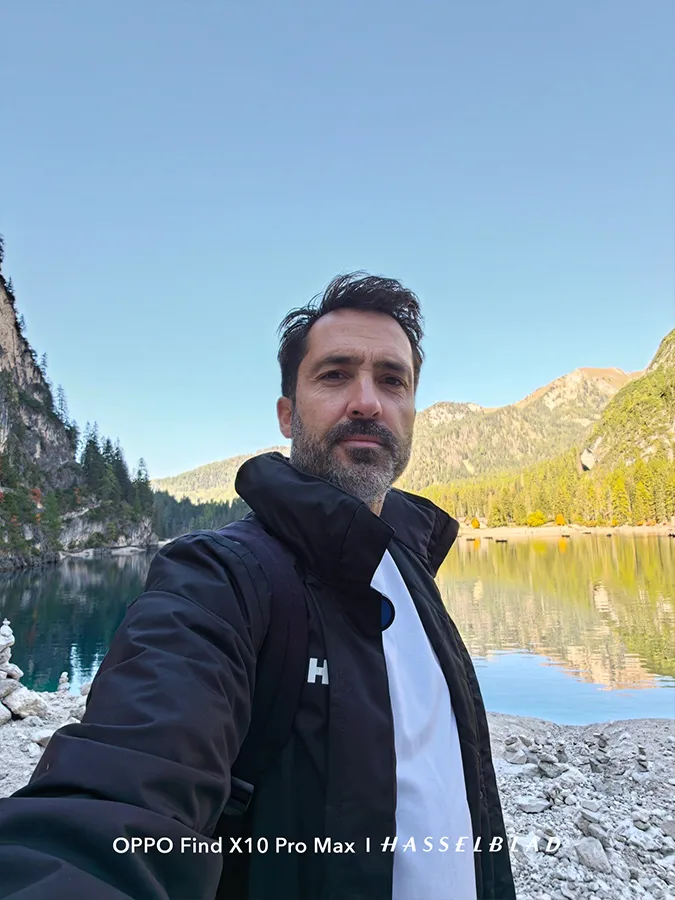 Gros plan d'un homme prenant un selfie dans un environnement naturel avec des montagnes, un lac et un ciel clair.