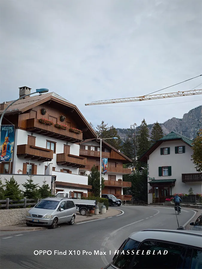 Rue d'un village alpin avec des maisons en bois, une voiture garée et des montagnes en arrière-plan sous un ciel nuageux.
