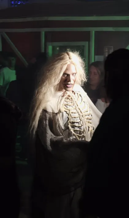 Personne déguisée avec une perruque blonde et une structure squelette sur la poitrine éclairée directement dans un environnement nocturne.
