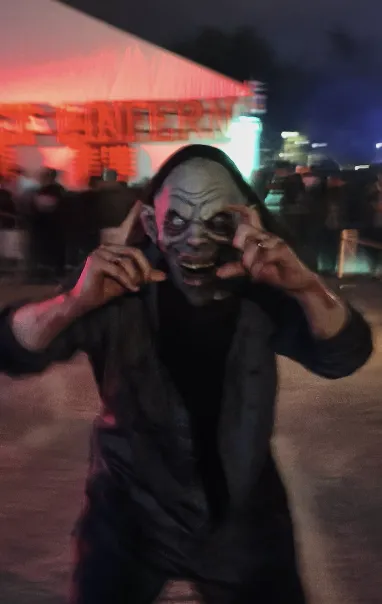 Personne portant un masque de créature effrayante faisant des gestes avec ses mains vers son visage lors d'un événement nocturne.