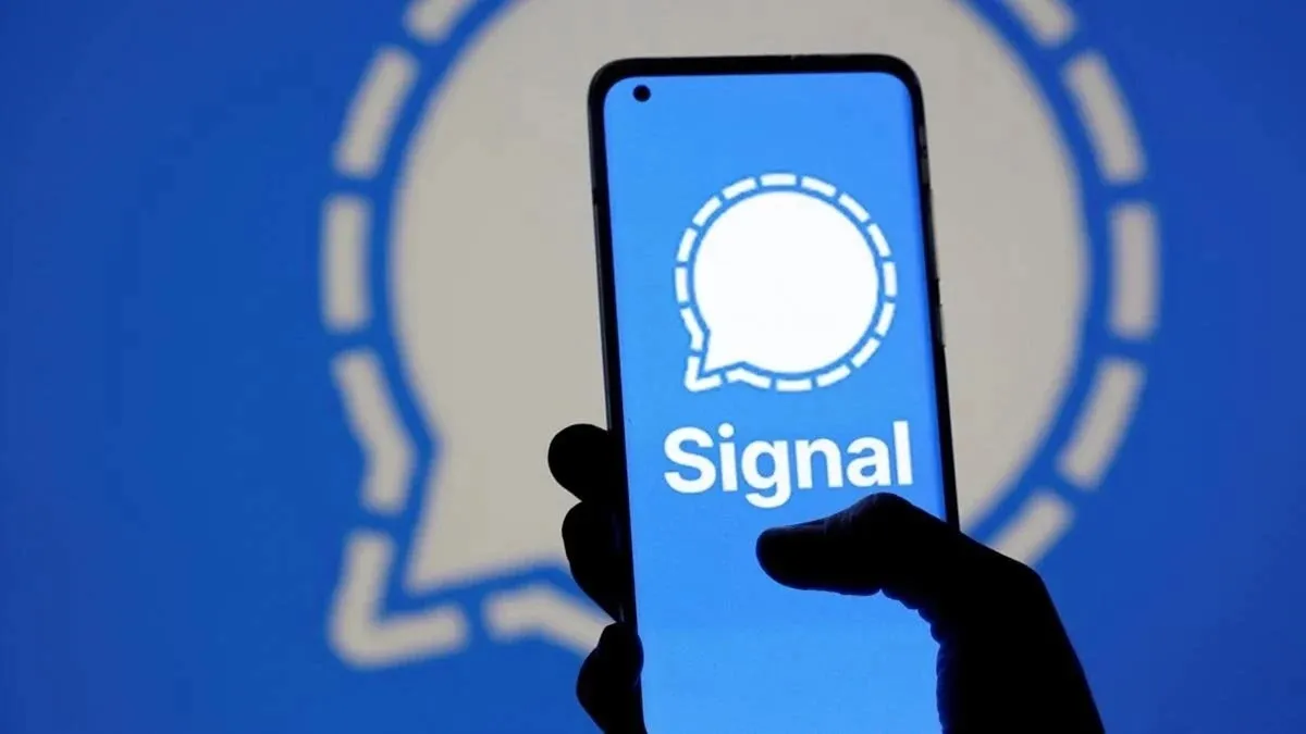 Main tenant un téléphone mobile avec le logo de l'application Signal à l'écran sur fond bleu.